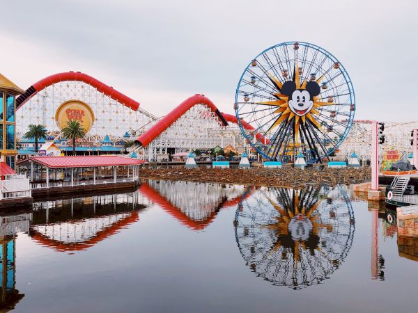 Amusement park with a large Ferris wheel and roller coaster, reflected in the water, featuring a familiar cartoon character’s face.