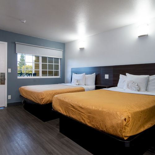 The image shows a hotel room with two beds, a window, and a closed door, featuring warm lighting and modern decor.