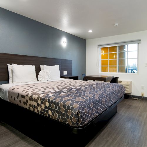 The image shows a hotel room with a large bed, hexagonal patterned bedding, nightstands, window, door, and minimal modern decor.