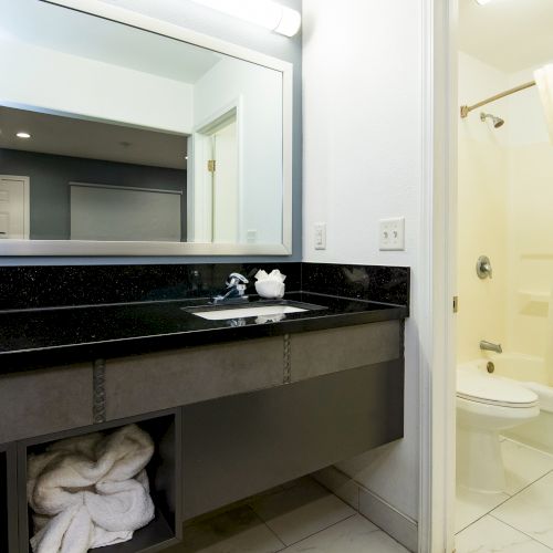 A clean, modern bathroom with a black countertop, large mirror, towels stored below, and a shower with a white curtain.