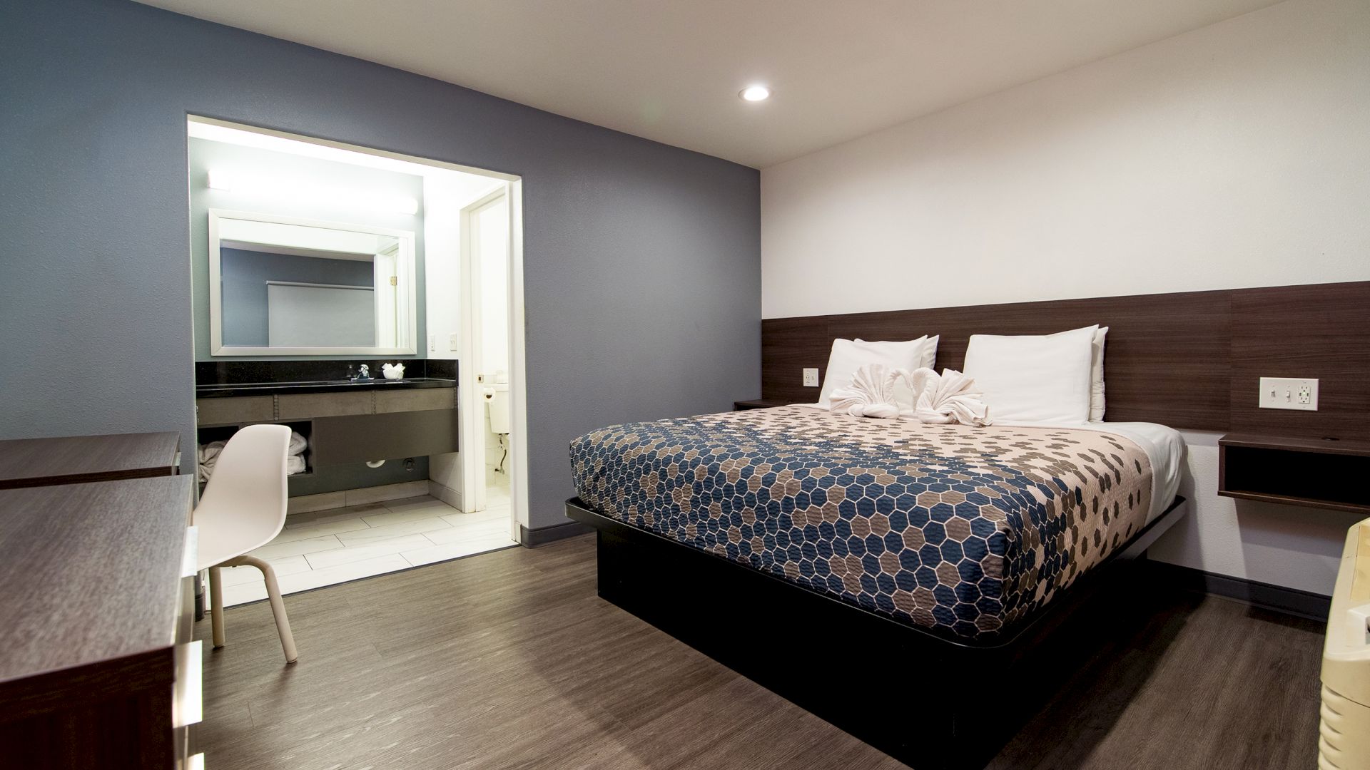 A modern hotel room with a bed, desk, chair, and bathroom area, featuring clean design and neutral colors.