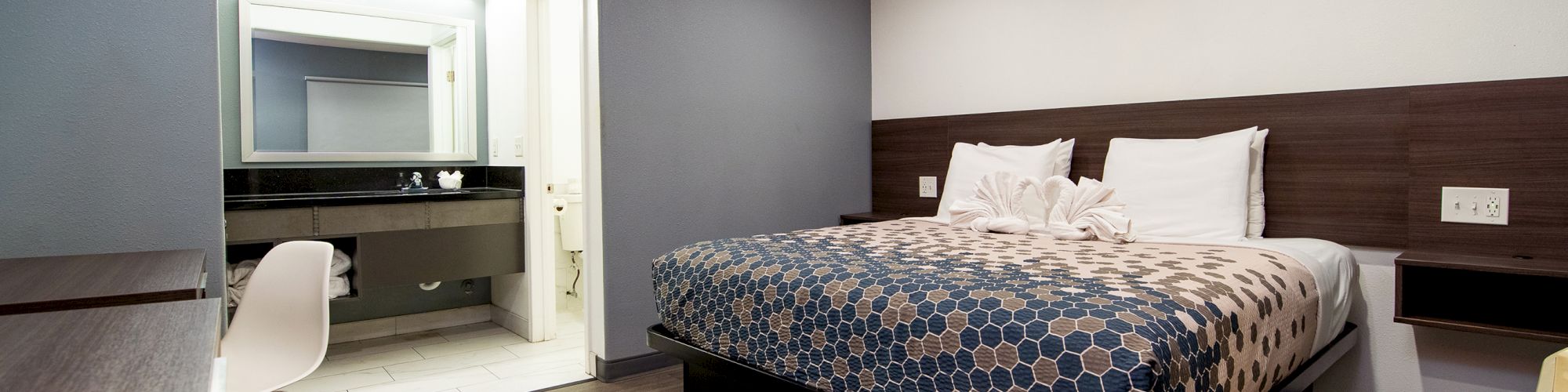 A modern hotel room with a bed, desk, chair, and bathroom area, featuring clean design and neutral colors.